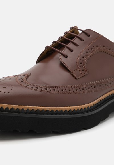 Brune lædersko med brogue-detaljer, rund tå, sort gummisål og kontrasterende tan mellemsål. Seks snørehuller til snørebånd.