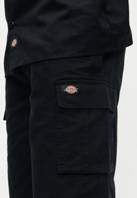 Pantalon cargo noir et chemise avec logos visibles de la marque Dickies sur l'ourlet de la chemise et le rabat de la poche cargo.