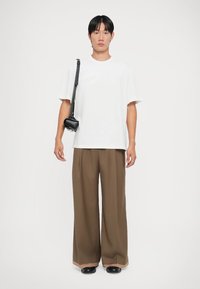 Witte T-shirt met korte mouwen, losse bruine broek en zwarte schoudertas. De broek heeft een wijdere snit en een gladde textuur.