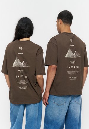 Dvaja ľudia nosia hnedé tričká s grafikou pyramídy a textom o púšti a večnosti, v kombinácii s voľnými modrými džínami.