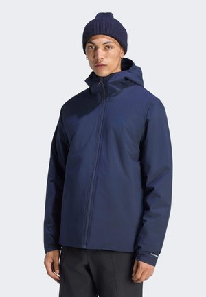 Junge Person trägt eine marineblaue Kapuzenjacke und eine passende gestrickte Mütze, schaut mit neutralem Gesichtsausdruck nach vorne.