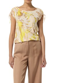 Kurzarmbluse aus leichtem Stoff mit beigem Hintergrund und gelben sowie braunen abstrakten Mustern. Geraffte Ärmel mit Bindebändern.