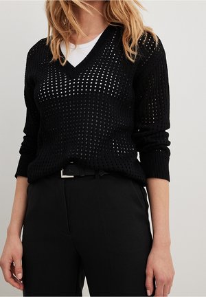 Kvinde iført sort perforeret V-hals sweater over hvid top, kombineret med sorte højttaljede bukser og bælte, stående mod en ensfarvet baggrund.