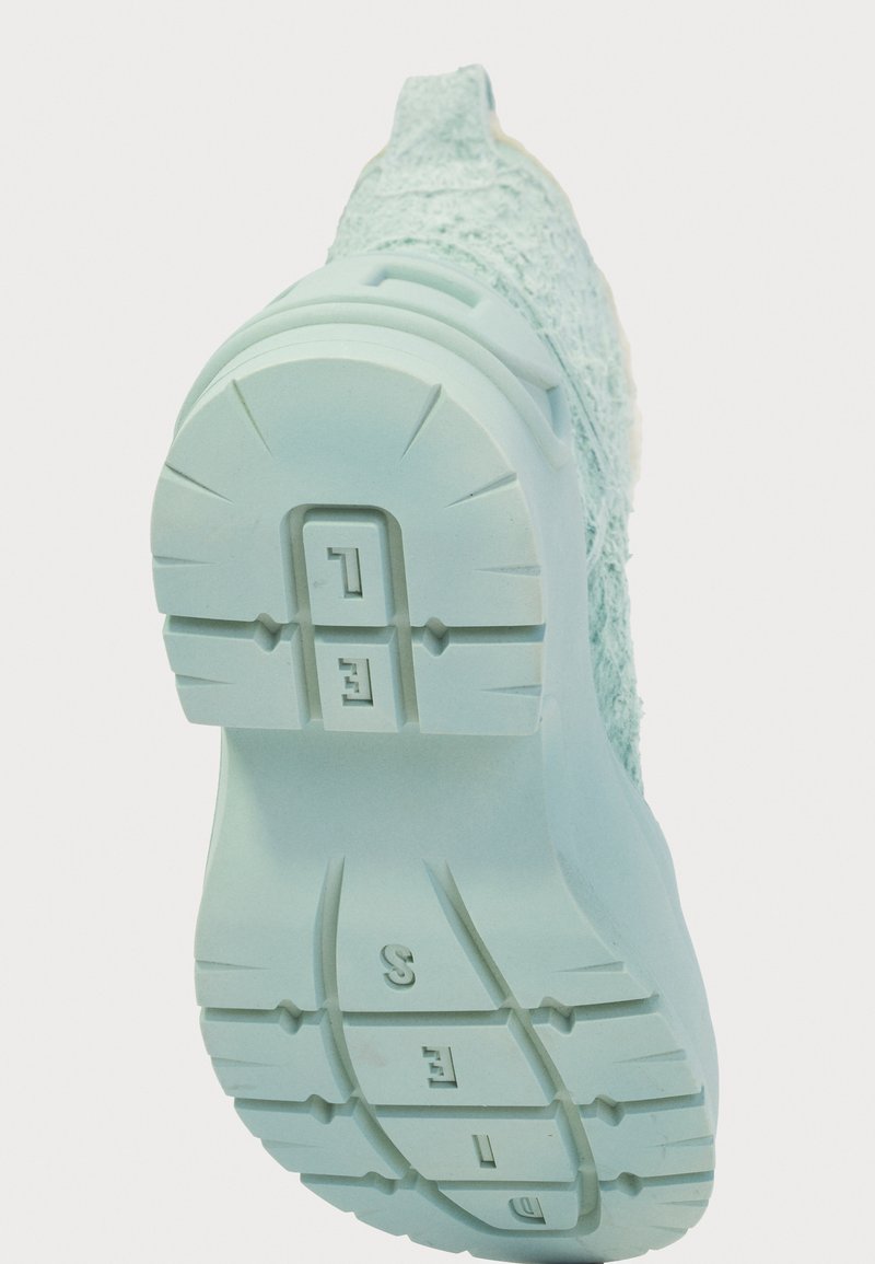 Helle blaue Sneaker-Sohle mit strukturiertem Profil und erhabenen Buchstaben und Zahlen, fotografiert von unten vor einem weißen Hintergrund.