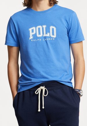 Osoba ubrana w niebieską koszulkę "POLO Ralph Lauren" i granatowe spodnie z sznurkiem, z rękami w kieszeniach i zegarkiem na lewym nadgarstku.