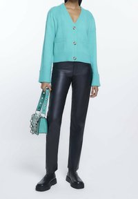 Tyrkysový pletený kardigan s V-výstřihem, dvěma předními kapsami a kovovými knoflíky; zkombinovaný s černými koženými kalhotami a silnými černými botami.