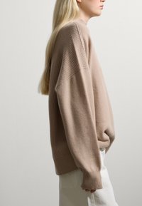 Beige übergroßer Pullover mit gerippter Textur, hohem Ausschnitt und fallenden Schultern, kombiniert mit weißen Hosen. Schlichtes, modernes Design.