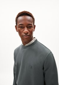 Sweatshirt gris avec un col rond et un logo brodé. Le tissu semble doux avec une texture lisse. Porté sur une chemise à col.