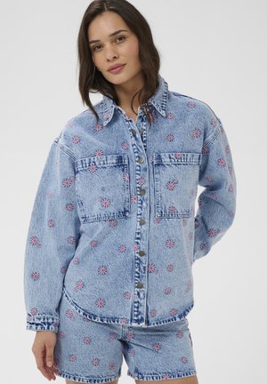 Kvinde iført lysblå denimjakke og shorts med lyserød blomsterbroderi, stående mod en ensfarvet lys baggrund.