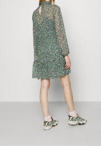 Vestido floral verde con mangas largas, espalda con abertura en forma de ojo de cerradura y dobladillo con volantes. Llevado con zapatillas estampadas y calcetines rosas.