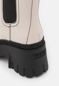 Semelle noire épaisse avec bord texturé et dessus en cuir beige comportant un panneau élastique noir et le logo Coach sur le talon.