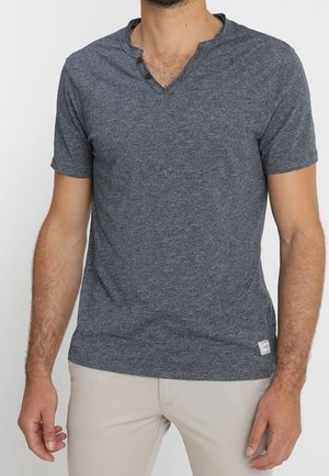 Mężczyzna ubrany w ciemnoszary krótki rękaw henley z dwoma guzikami i jasnobeżowe spodnie, stojący na jednolitym tle.