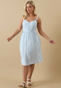 Ljust blå blommig klänning med tunna axelband, V-ringning, knappdetaljer framtill och en gathered kjol. Ett par vita sandaler synliga.