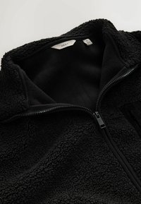 Černá fleecová bunda s vysokým límcem a zipem vpředu. Má měkkou, strukturovanou látku a uvnitř oblasti krku je štítek velikosti M.