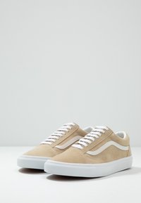 Sneakers in suede beige con laccetti bianchi e una striscia laterale bianca. Presentano una suola in gomma piatta e una punta rotonda. Il materiale testurizzato crea una finitura morbida.