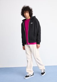Black zip-up hoodie, pink t-shirt underneath, cream striped pants, and black sneakers. Curly hair, standing pose on plain background.