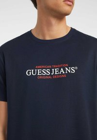 Tamsiai mėlynos spalvos medvilninė marškinėliai su centriniu grafiniu elementu: "AMERICAN TRADITION GUESS JEANS ORIGINAL DESIGNS" baltos ir raudonos spalvos tekstu.
