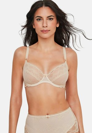 Femme aux longs cheveux foncés portant un soutien-gorge en dentelle beige et une culotte taille haute assortie en dentelle, se tenant devant un fond blanc uni.