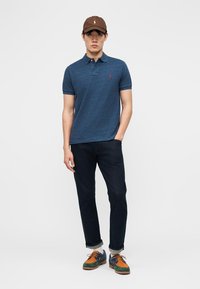 Mladý muž v hnedej čiapke, modrej polokošeli, tmavých džínsach vyhrnutých nad členky a zeleno-oranžových šnurovacích topánkach, stojaci s jednou rukou v vrecku.