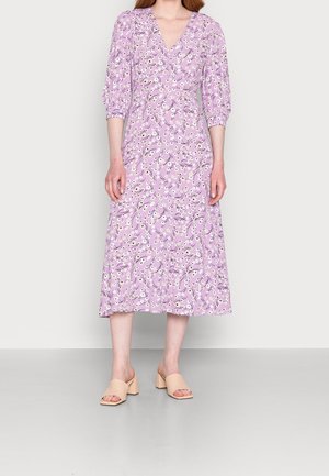Femme portant une robe midi lavande à imprimé floral blanc, manches trois-quarts bouffantes, et des sandales beiges à talons blocs ouvertes aux orteils.