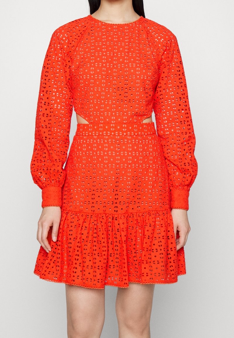 Femme portant une robe en dentelle ajourée orange vif avec des manches longues bouffantes, une taille découpée et un ourlet volanté au-dessus du genou.