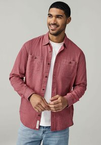 Chemise à manches longues à motifs rouges en tissu tissé, avec un devant boutonné, deux poches poitrine et un t-shirt à col rond blanc en dessous.