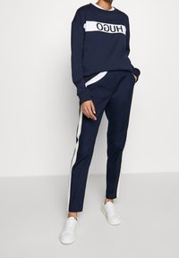 Sudadera azul marino con panel blanco y logo "HUGO", combinada con pantalones azul marino con franjas blancas laterales. Zapatillas blancas completan el conjunto.