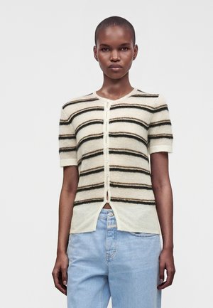 Vrouw draagt een crèmekleurige trui met korte mouwen en knopen, met zwarte en bruine horizontale strepen en lichtblauwe high-waisted jeans.
