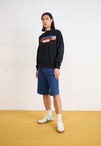 Sudadera negra con gráfico y texto colorido, combinada con pantalones cortos de mezclilla. Zapatillas blancas con acentos verdes. Fondo texturizado.