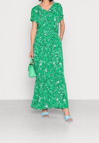 Nainen, jolla on päällään vihreä kukkakuosisella maxi-mekolla, jossa on lyhyet hihat, hän pitää kädessään vihreää käsilaukkua ja jalassaan on vaaleansinisiä avokkaita.