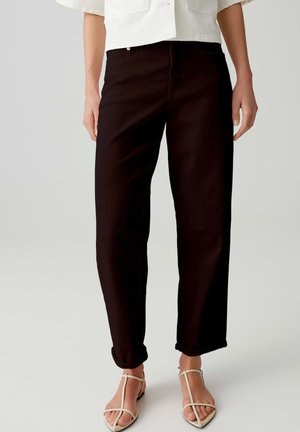 Persona che indossa pantaloni neri a gamba dritta risvoltati alle caviglie e sandali beige con punta aperta, in piedi su uno sfondo chiaro.