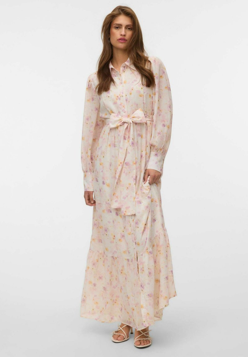 Femme portant une longue robe fleurie rose clair avec des manches longues, un col et un nœud à la taille, associée à des sandales blanches à brides.