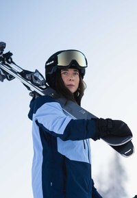 Ski equipment with gray skis and blue-and-black insulated jacket. Black helmet and mirrored goggles, holding skis over shoulder on snow.