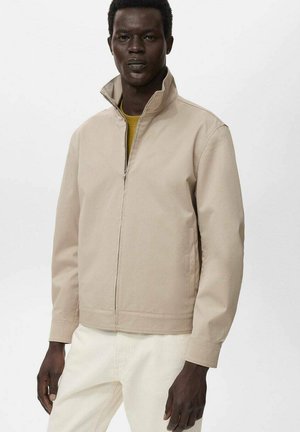 Beige zip-up jakke med høj krave, der har en glat tekstur og minimalistisk design. Bæret over en gul skjorte og lyse bukser.