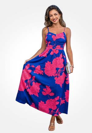 Kobieta uśmiechająca się, ubrana w niebieską suknię maxi z jasnoróżowymi wzorami kwiatowymi, trzyma suknię jedną ręką, a w drugiej kopertówkę.