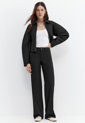 Nő fehér fal előtt áll, fekete cipzáras dzsekit, széles szárú fekete nadrágot, fehér crop topot és bézs cipőt visel, egyik kezét a csípőjén tartva.