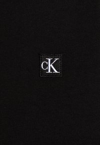 Schwarzer Strickstoff mit einem weißen, gestickten 'cK'-Logo auf einem quadratischen Patch. Die Textur ist glatt mit einem gerippten Muster.