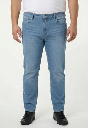 Mann trägt hellblaue Slim-Fit-Jeans mit fünf Taschen und schwarze Turnschuhe, steht vor einem schlichten Hintergrund.