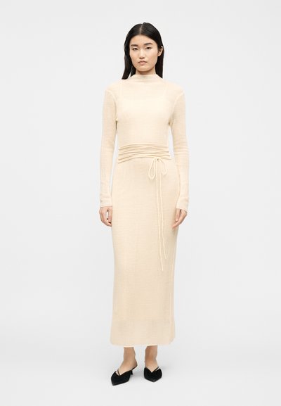 Femme aux longs cheveux noirs portant une robe longue à manches longues en maille côtelée crème avec des liens à la taille et des escarpins noirs à bouts pointus, debout devant un fond blanc.