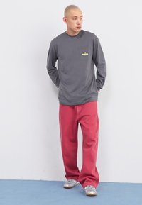Gray long-sleeve shirt with a front pocket and colorful graphic, paired with red pants and gray sneakers on a light blue floor.