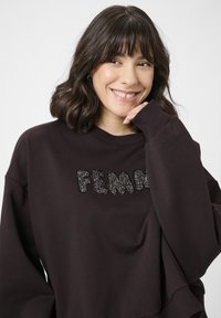 Usmievajúca sa žena s vlnitými tmavými vlasmi do ramien má na sebe tmavohnedú mikinu s čiernym korálkovým textom "FEMME", opiera si bradu o ruku.