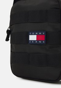 Borsa in tessuto nera con cinturino orizzontale, un logo rettangolare con elementi rossi, bianchi e blu navy, e chiusura a zip.