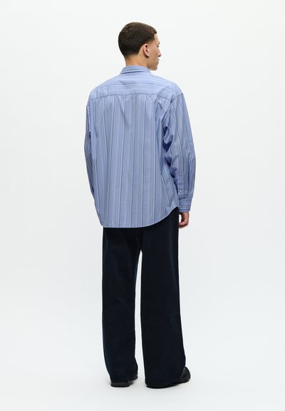Camisa de rayas azul clara con mangas largas, puños abotonados y un corte holgado, combinada con pantalones de pana anchos negros.