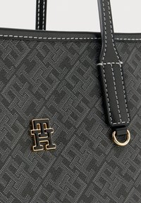 Svart teksturert håndveske med hvite sømmer, med et gull- og svart sammenflettet "TH"-logo og lærstropp med gullring.