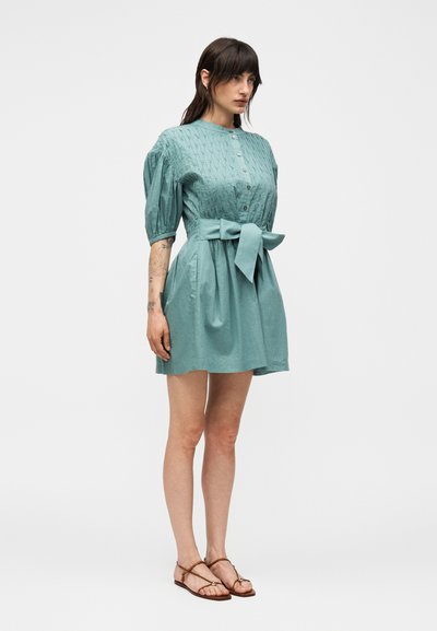 Γυναίκα που στέκεται, φοράει κοντό τιρκουάζ φόρεμα με φουσκωτά μανίκια, κλείσιμο με κουμπιά μπροστά, ζώνη στη μέση και καφέ σανδάλια με λουράκια και επίπεδη σόλα.