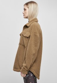 Brun fleecejakke med kragehals, to frontlommer og avslappet passform. Myk tekstur med noe oversized ermer.