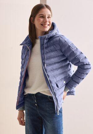 Doudoune bleu clair avec fermeture éclair, capuche et intérieur côtelé. Présente des coutures verticales et des poches latérales, portée par-dessus un léger pull.
