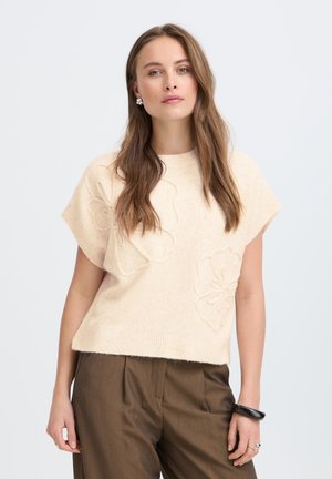 Femme aux longs cheveux bruns portant un pull crème à manches courtes avec des motifs floraux et un pantalon marron, sur un fond clair uni.
