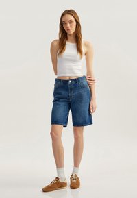 Bermudas de mezclilla en un lavado azul medio, con una cintura de altura media, diseño tradicional de cinco bolsillos y dobladillos acabados. Combinadas con una camiseta sin mangas blanca.