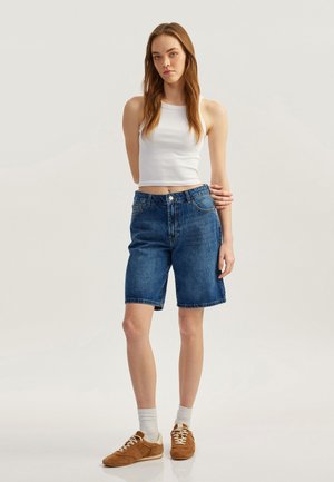 Bermudas de mezclilla en un lavado azul medio, con una cintura de altura media, diseño tradicional de cinco bolsillos y dobladillos acabados. Combinadas con una camiseta sin mangas blanca.